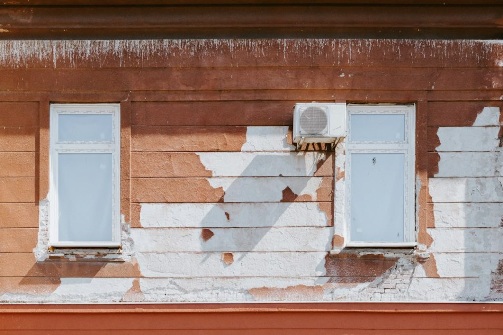 Two windows on a weathered red wall with peeling white paint and an air conditioner